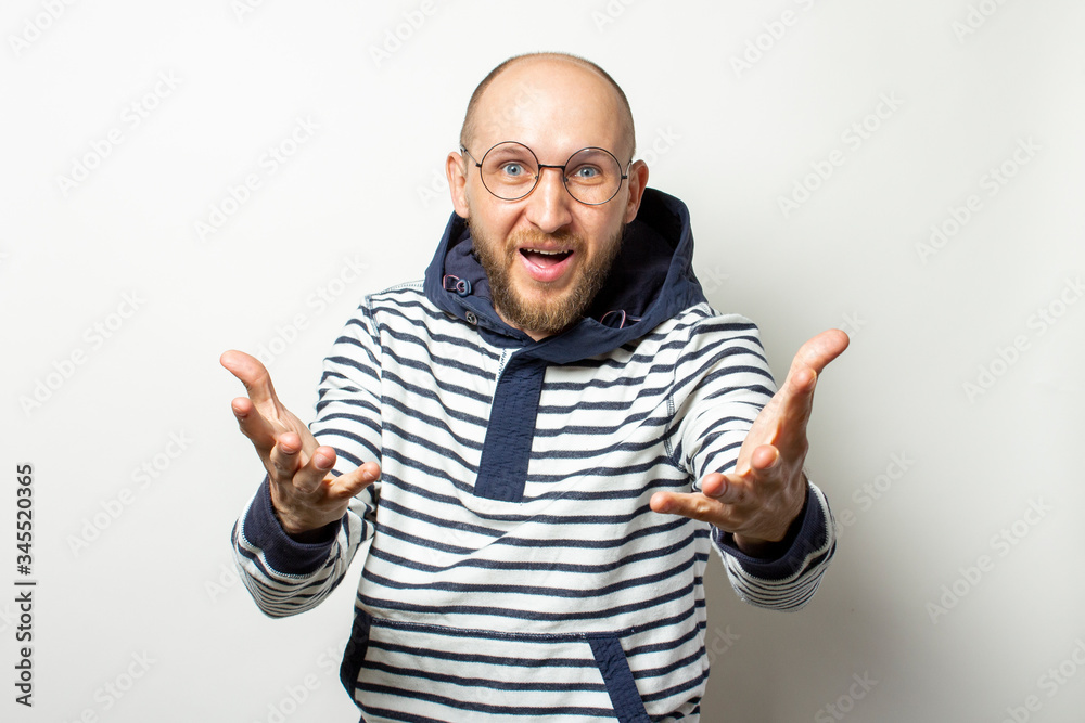 Portrait of a Bald Young man with a beard in glasses, a sweater with a hood, pulls his hands to the viewer on an isolated light background. Emotional face. Gesture invitation, call