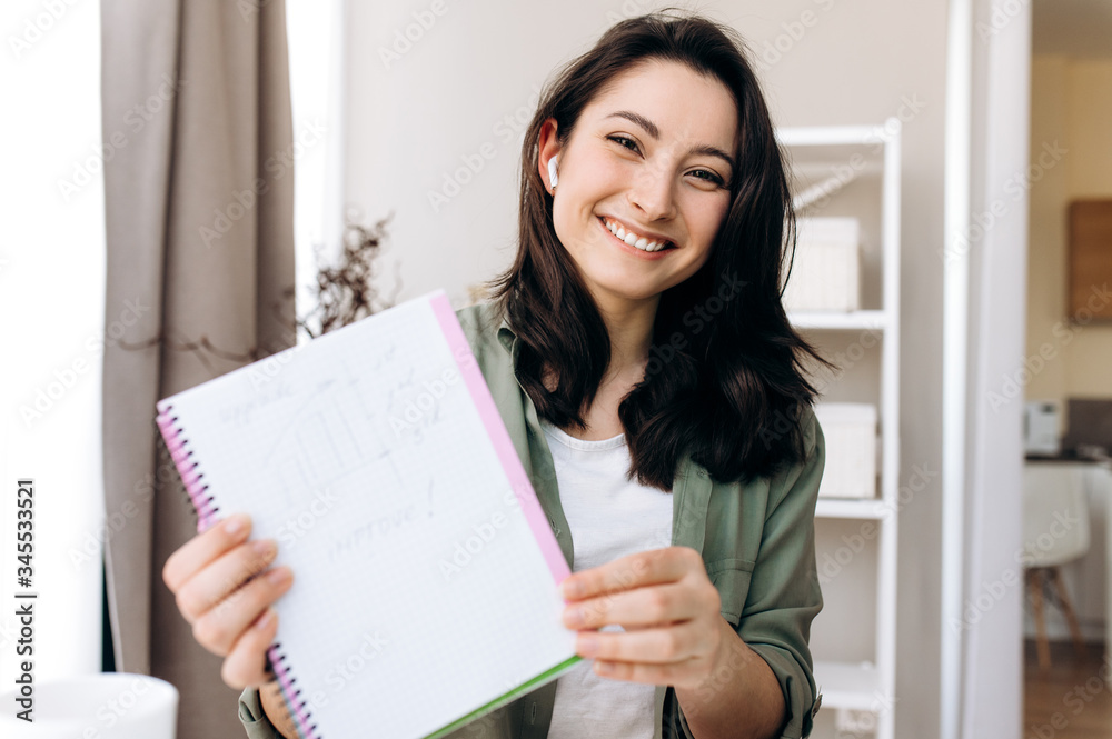 Attractive smiling girl shows a notebook while talking on a video conference with colleagues using a laptop. Remotely work Attractive smiling girl shows a notebook while talking on a video conference with colleagues using a laptop. Remotely work
