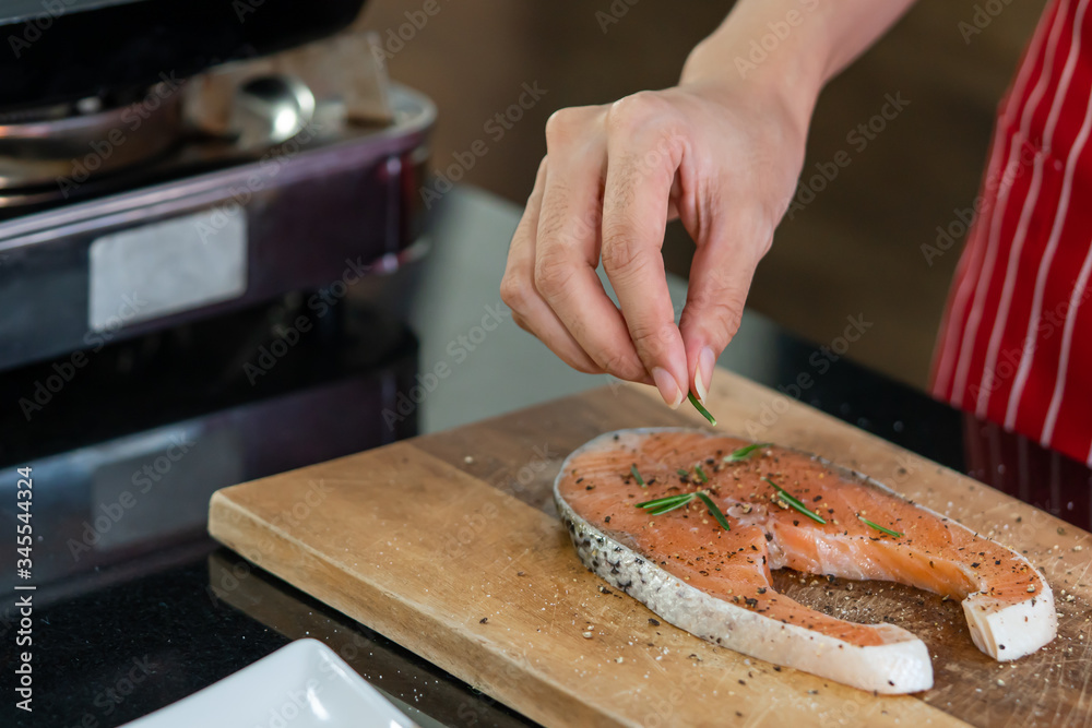 Hand of woman is sprinkled with pepper and rosemary on a piece of salmon cooking delicious salmon steak at the kitchen.