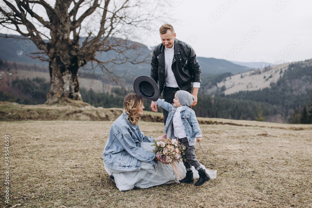 Fototapeta premium Stylish couple in the mountains. Stock Photo. A guy in a leather jacket and a young girl in a gray-blue wedding dress and a denim jacket with their son on the background of a large tree and sky