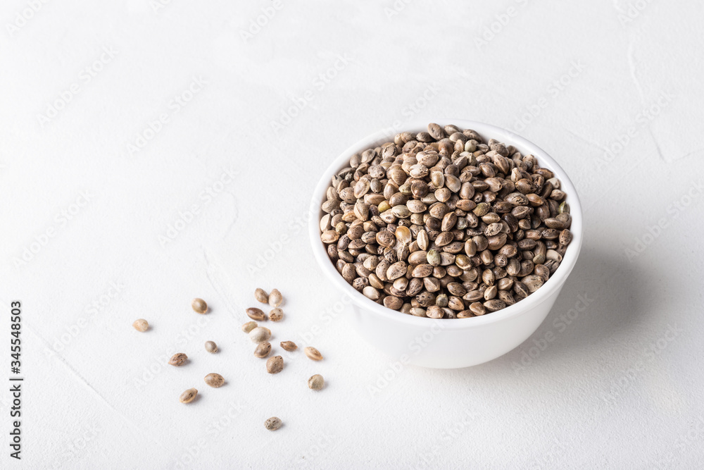 Raw organic unrefined hemp seeds in small white bowl on white concrete background. Raw organic unrefined hemp seeds in small white bowl on white concrete background.
