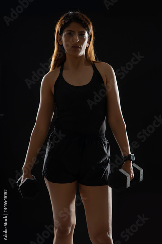 Woman standing with dumbbells in her hand in front of a dark background. 