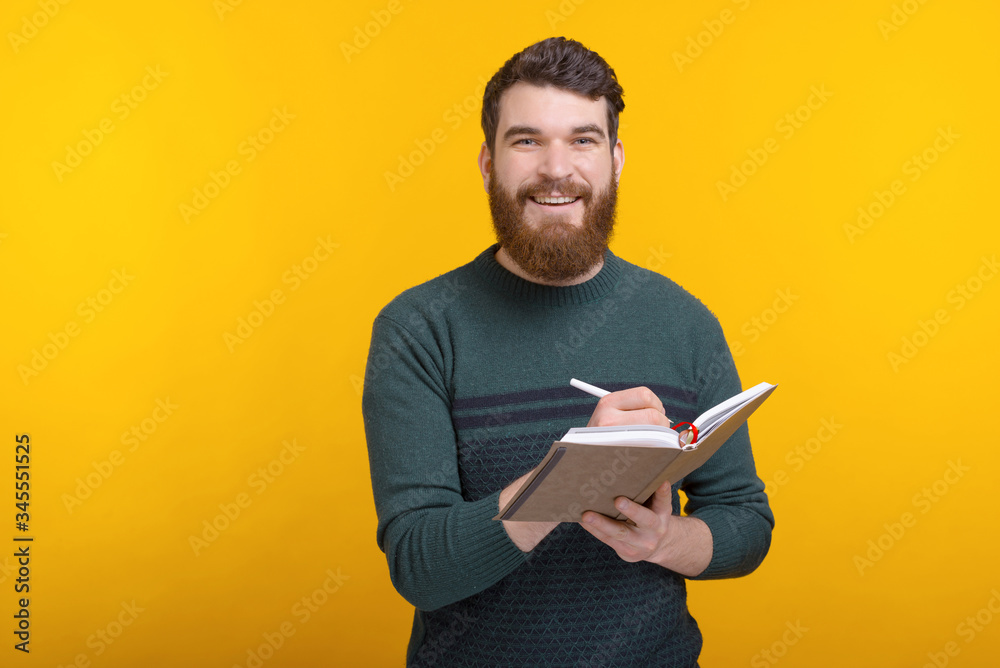 Bearded man looking at the camera is writing someting in his journal on yellow background. Bearded man looking at the camera is writing someting in his journal on yellow background.