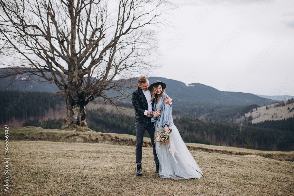 Stylish couple in the mountains. A guy in a leather jacket and a young girl in a gray-blue wedding dress and a denim jacket are going together on the background of a large tree, sky and landscapes