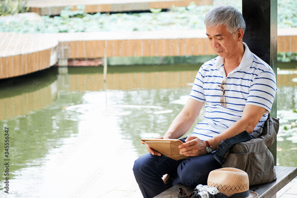 Aged Chinese man sitting by pond in city park and reading news or watching interesting video on tablet computer