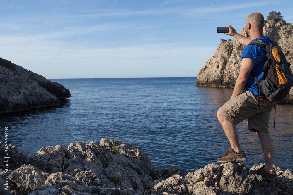 A young and bald man taking photographs with the mobile to a seascape on a beach without people in Mallorca. The man is wearing a backpack and it is early in the morning
