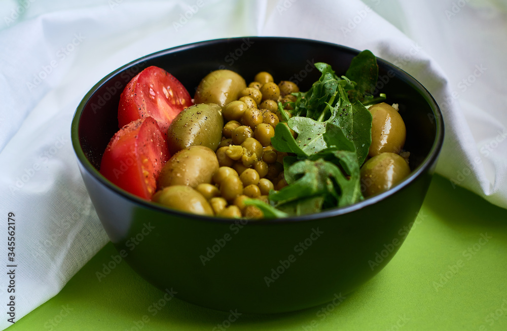 Green summer background and white cloth with  black bowl and delicious useful breakfast of porridge and vegetables. Tomatoes, olives, arugula, peas.