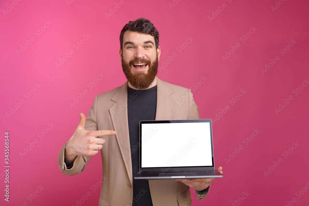 Excited bearded man smiling at the camera is pointing at his laptop he is holding on pink background.