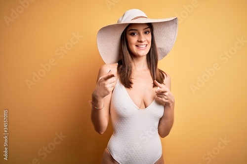 Young beautiful brunette woman on vacation wearing swimsuit and summer hat pointing fingers to camera with happy and funny face. Good energy and vibes.