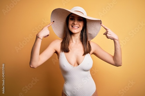 Young beautiful brunette woman on vacation wearing swimsuit and summer hat smiling pointing to head with both hands finger, great idea or thought, good memory