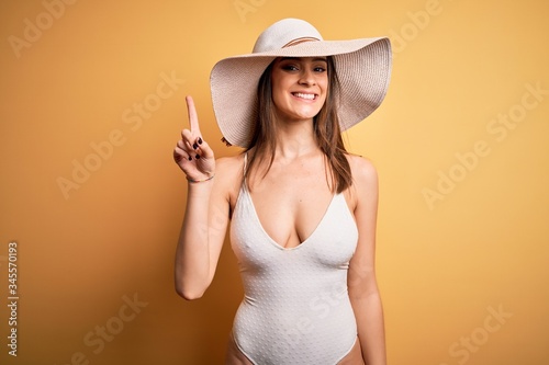 Young beautiful brunette woman on vacation wearing swimsuit and summer hat showing and pointing up with finger number one while smiling confident and happy.