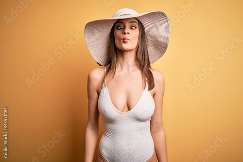 Young beautiful brunette woman on vacation wearing swimsuit and summer hat making fish face with lips, crazy and comical gesture. Funny expression.