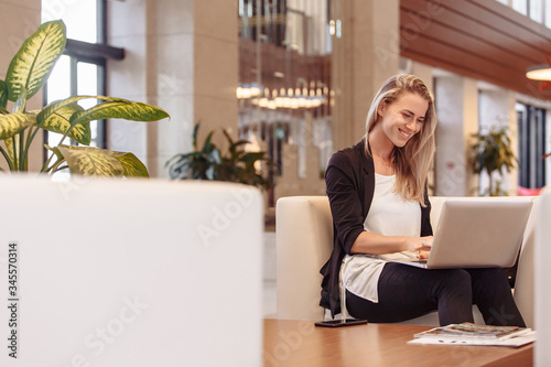 Laughing young beautiful blonde woman is sitting in lobby of hotel and waiting for check-in checking mail and social networks using a laptop and high-speed Internet. Successful beautiful woman concept
