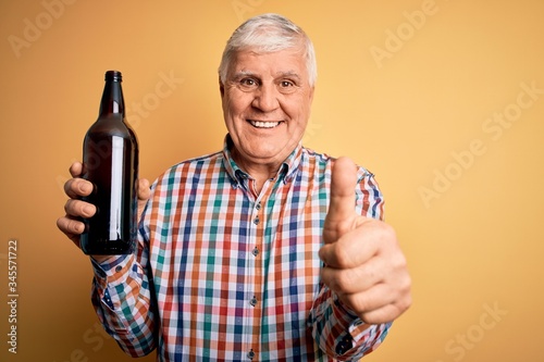 Fotografia Senior handsome hoary man drinking bottle of beer standing over isolated yellow