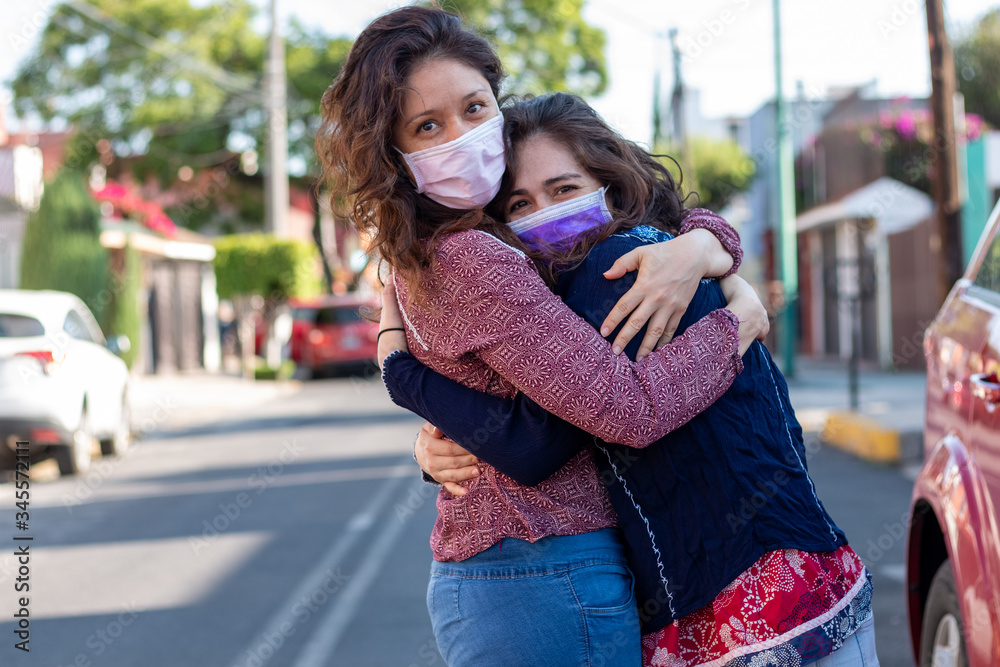 two young Latinas wearing protective medical masks, dressed in casual clothing, standing and hugging and looking at each other. Coronavirus, pandemic,