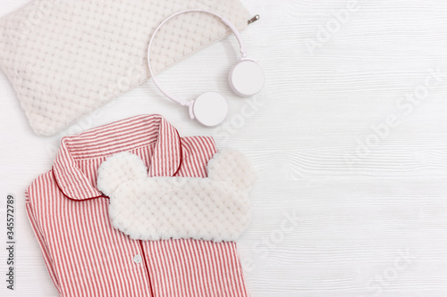 Pink pajamas for Girls, eye mask for sleeping, headphones, soft cushion on white wood. Comfortable nightwear for peaceful sleep. Top view. Flat lay.
