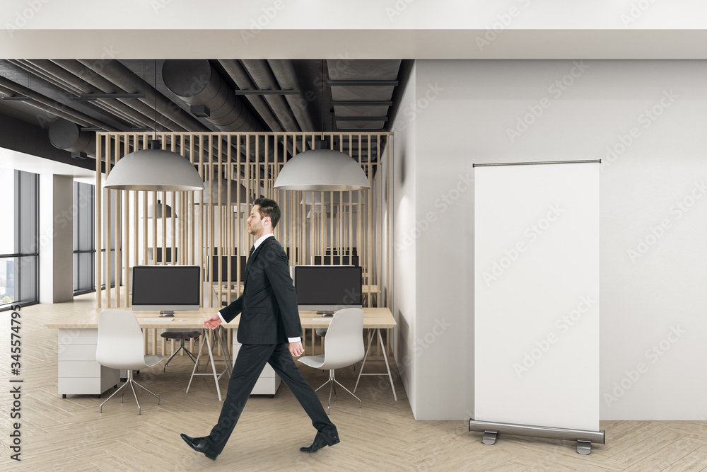 Businessman walking in modern office with computers and vertical poster on wall