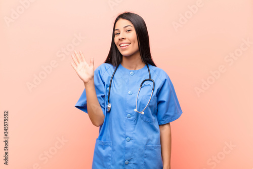 young latin nurse smiling happily and cheerfully, waving hand, welcoming and greeting you, or saying goodbye against pink wall photo