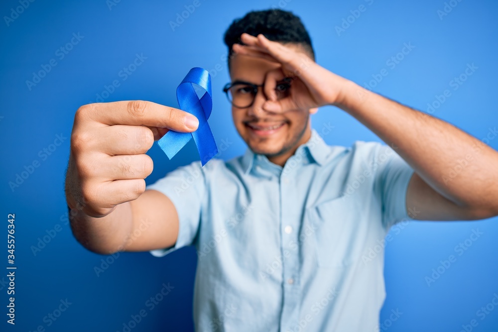 Young handsome man holding blue cancer ribbon standing over isolated background with happy face smiling doing ok sign with hand on eye looking through fingers