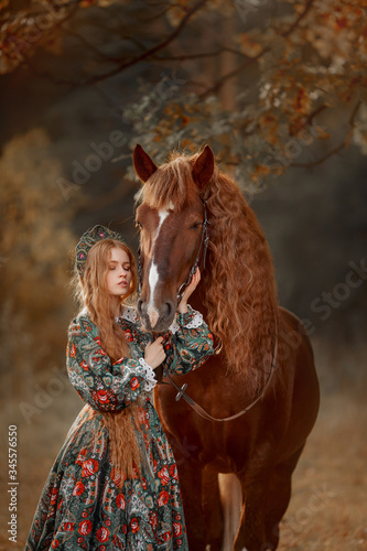 Beautiful long-haired blonde young woman in national russian style with red Vladimir draft horse in autumn forest © Julia Shepeleva