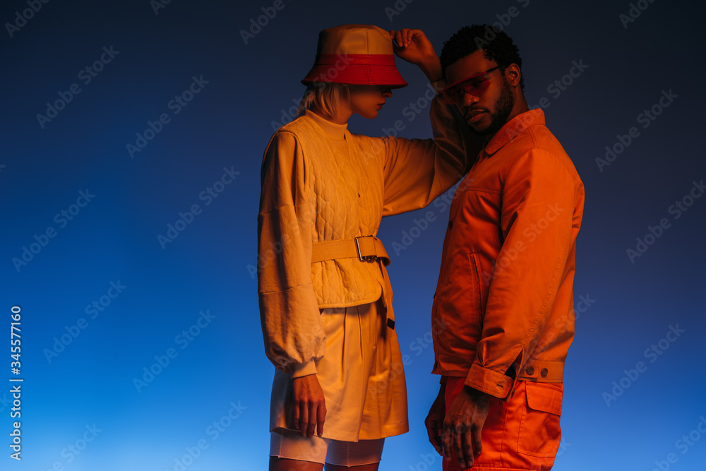 fashionable interracial couple in futuristic clothes, sunglasses and hat posing on blue in orange light