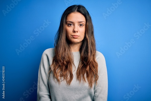 Young beautiful brunette woman wearing casual sweater standing over blue background with serious expression on face. Simple and natural looking at the camera.