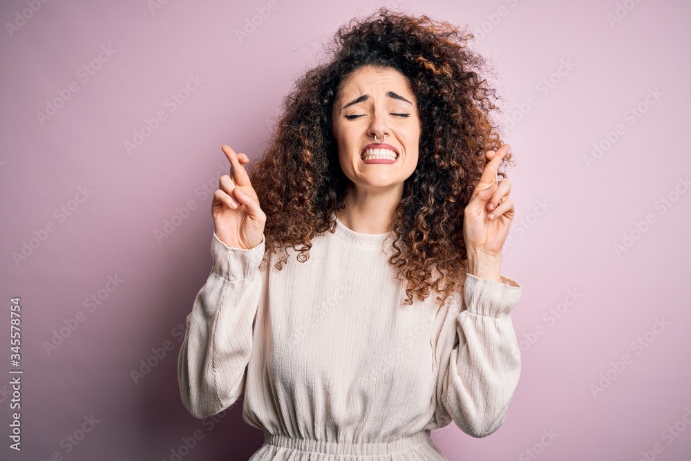 Beautiful woman with curly hair and piercing wearing casual sweater over pink background gesturing finger crossed smiling with hope and eyes closed. Luck and superstitious concept. Beautiful woman with curly hair and piercing wearing casual sweater over pink background gesturing finger crossed smiling with hope and eyes closed. Luck and superstitious concept.