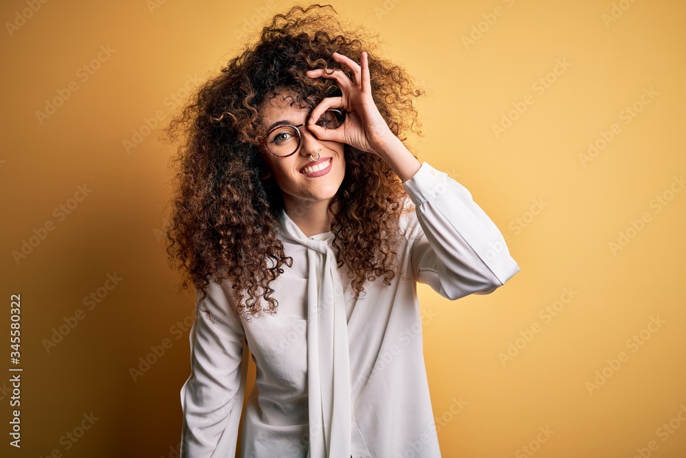 Young beautiful brunette woman with curly hair and piercing wearing shirt and glasses doing ok gesture with hand smiling, eye looking through fingers with happy face. Young beautiful brunette woman with curly hair and piercing wearing shirt and glasses doing ok gesture with hand smiling, eye looking through fingers with happy face.