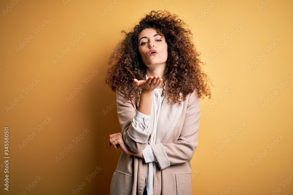 Young beautiful brunette woman with curly hair and piercing wearing casual t-shirt and diadem looking at the camera blowing a kiss with hand on air being lovely and sexy. Love expression. Young beautiful brunette woman with curly hair and piercing wearing casual t-shirt and diadem looking at the camera blowing a kiss with hand on air being lovely and sexy. Love expression.