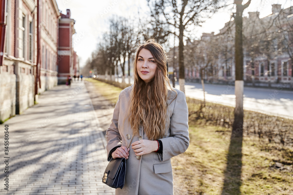 portrait of a blonde with long hair in a light coat with a bag. On a street with trees and an old red building portrait of a blonde with long hair in a light coat with a bag. On a street with trees and an old red building