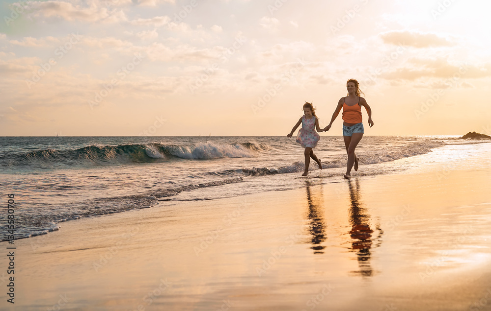 Happy loving family mother and daughter running on the beach at sunset - Mum having fun with her kid long sea shore during summer holidays - Parent vacation time lifestyle concept