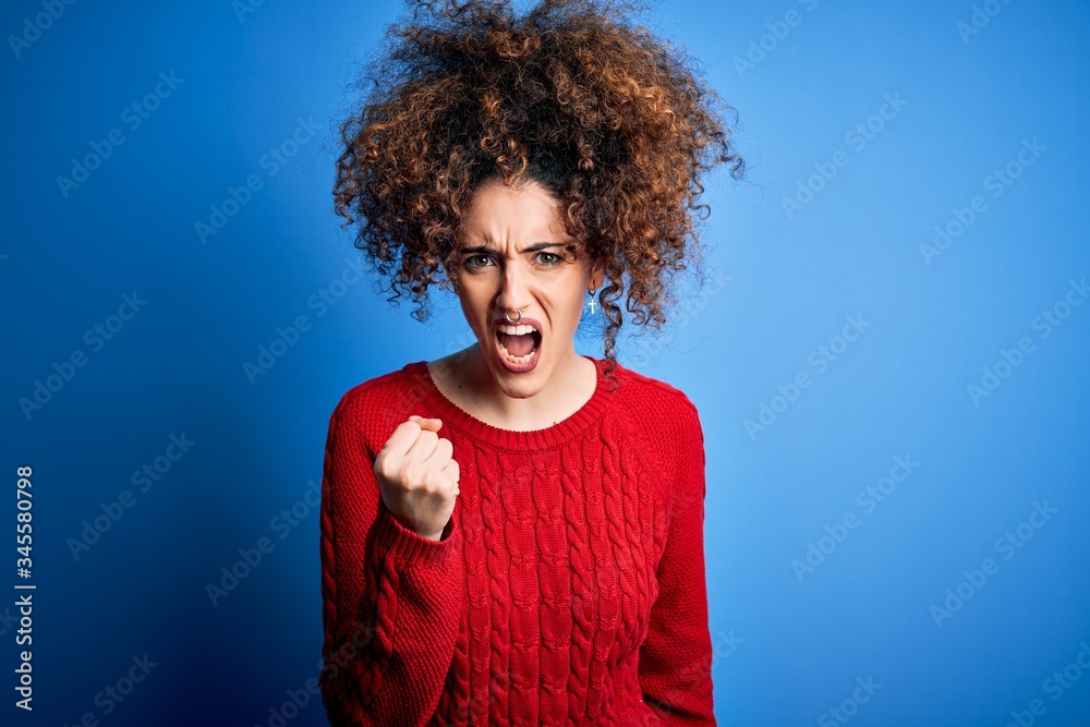 Young beautiful woman with curly hair and piercing wearing casual red sweater angry and mad raising fist frustrated and furious while shouting with anger. Rage and aggressive concept. Young beautiful woman with curly hair and piercing wearing casual red sweater angry and mad raising fist frustrated and furious while shouting with anger. Rage and aggressive concept.