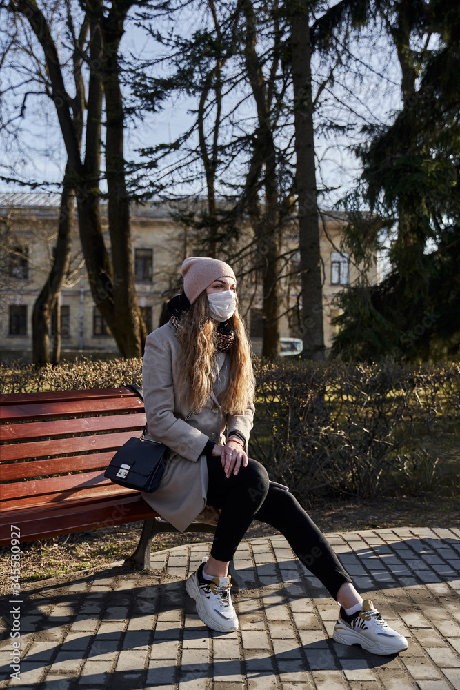 The blonde in a hat and coat in white shoes with a bag in a medical protective mask sits on a bench in the park. she basks in the sun