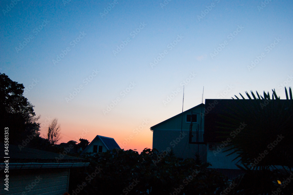 Sunset in the tropics - a dark blue sky with a strip of red setting sun against the background of houses, plants and palm trees. Sunset in the tropics - a dark blue sky with a strip of red setting sun against the background of houses, plants and palm trees.