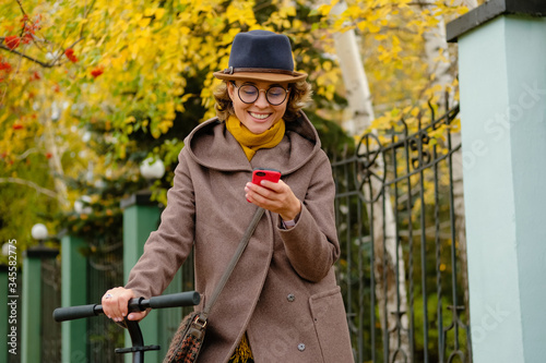 Beautiful girl in hat and glasses riding a scooter on a background of autumn. A modern young woman is surfing in the Internet, social networks, online orders