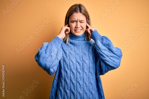 Young beautiful blonde woman wearing turtleneck sweater over yellow isolated background covering ears with fingers with annoyed expression for the noise of loud music. Deaf concept.