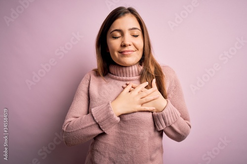 Beautiful young woman wearing turtleneck sweater over pink isolated background smiling with hands on chest with closed eyes and grateful gesture on face. Health concept.