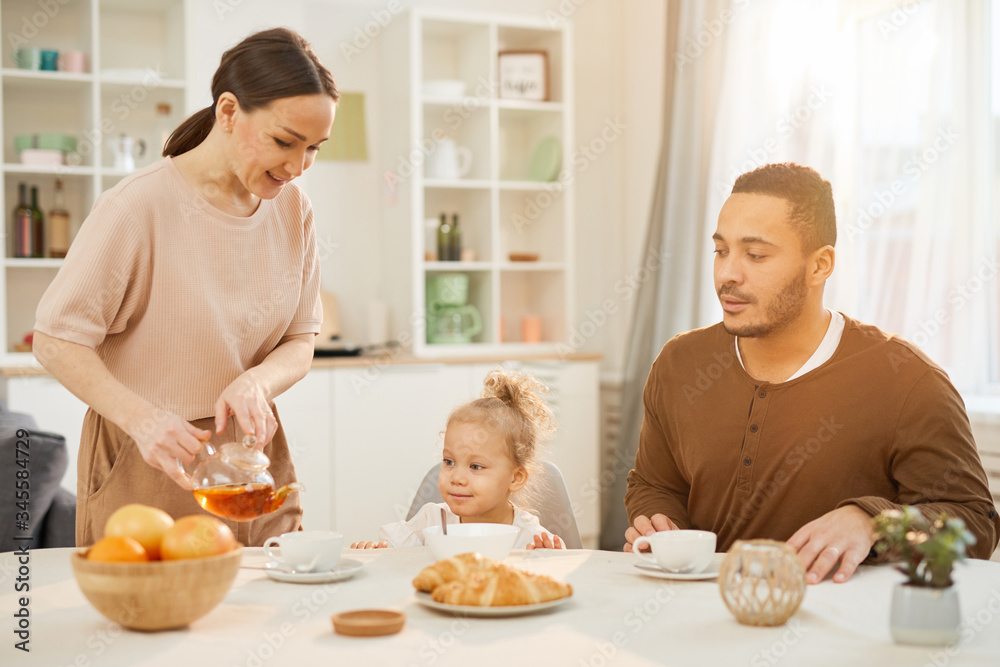 Young woman pouring tea into cups for her little daughter and husband during breakfast time at home