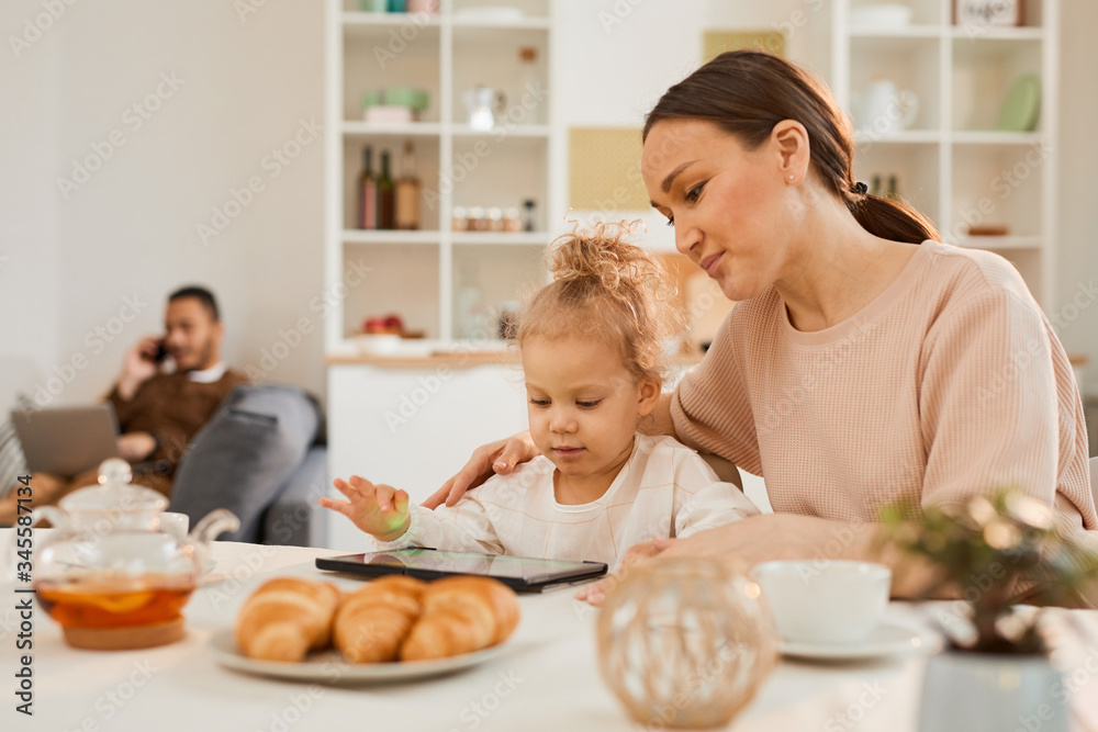 Loving mother and her cute little daughter sitting at table watching educational videos on digital tablet Loving mother and her cute little daughter sitting at table watching educational videos on digital tablet