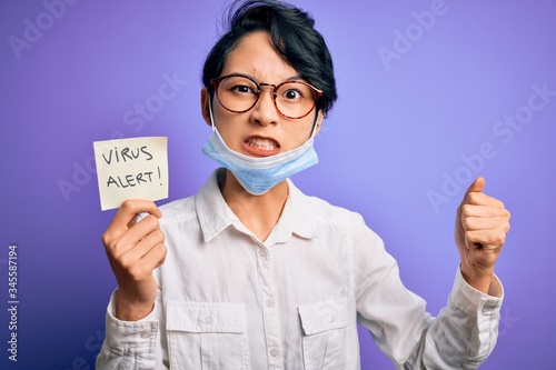Young beautiful asian girl wearing medical mask holding reminder with virus alert message annoyed and frustrated shouting with anger  crazy and yelling with raised hand  anger concept