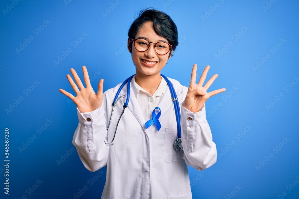 Young beautiful asian doctor girl wearing stethoscope and coat with blue cancer ribbon showing and pointing up with fingers number nine while smiling confident and happy. Young beautiful asian doctor girl wearing stethoscope and coat with blue cancer ribbon showing and pointing up with fingers number nine while smiling confident and happy.