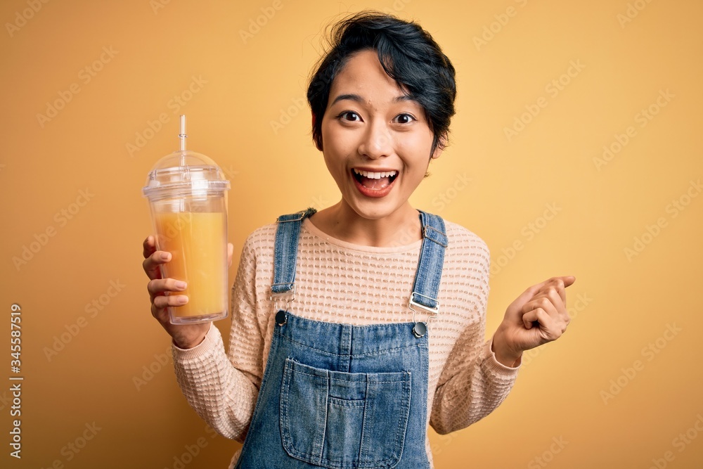 Young beautiful chinese woman drinking healthy orange juice over isolated yellow background screaming proud and celebrating victory and success very excited, cheering emotion Young beautiful chinese woman drinking healthy orange juice over isolated yellow background screaming proud and celebrating victory and success very excited, cheering emotion