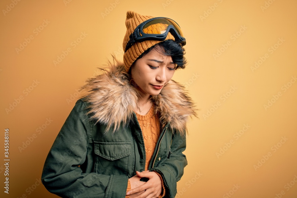 Beautiful asian skier girl wearing snow sportswear using ski goggles over yellow background with hand on stomach because indigestion, painful illness feeling unwell. Ache concept. Beautiful asian skier girl wearing snow sportswear using ski goggles over yellow background with hand on stomach because indigestion, painful illness feeling unwell. Ache concept.