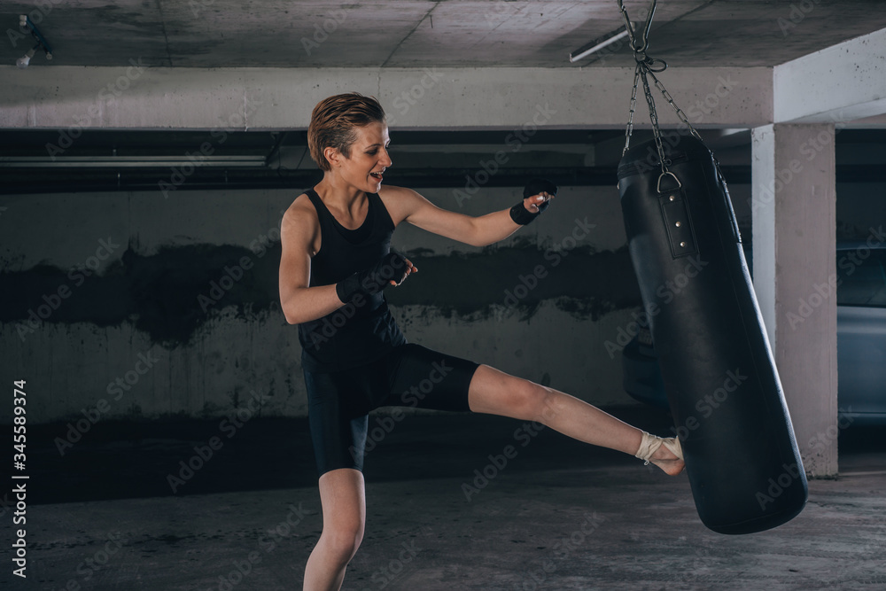 Caucasian woman in sportswear and with black bandages kicking the boxing bag inside a garage