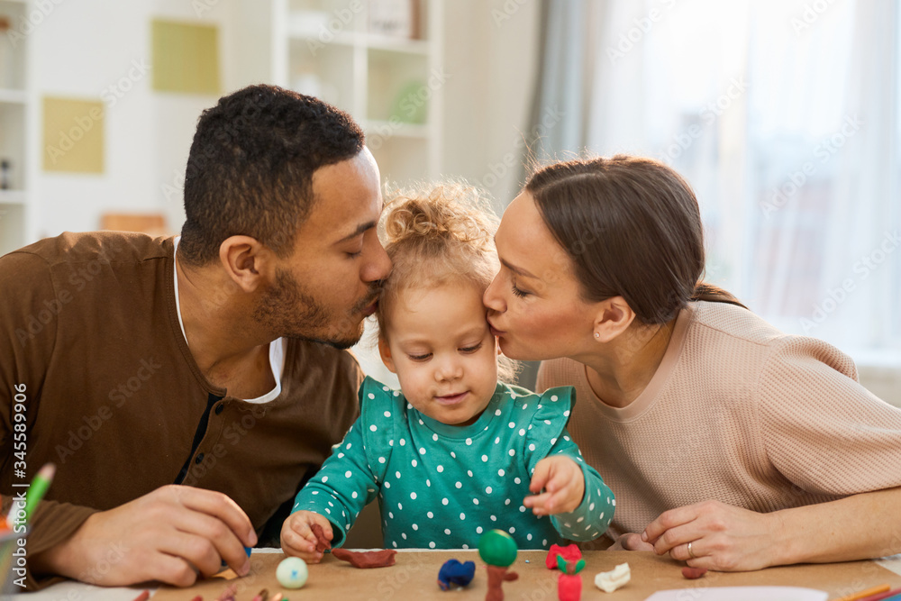 Obraz premium Young father and mother kissing their cute little daughter while she is making play dough shapes, horizontal shot