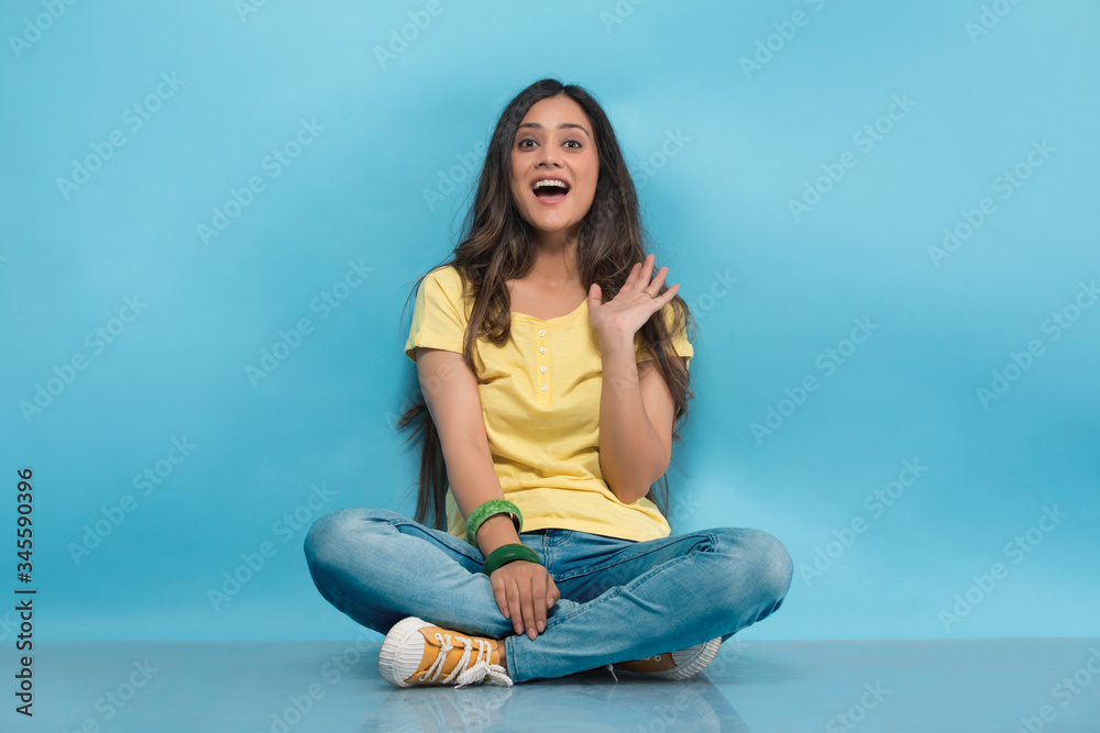 Happy teenage girl in jeans and t-shirt sitting cross legged on the floor and waving her hand
Happy teenage girl in jeans and t-shirt sitting cross legged on the floor and waving her hand