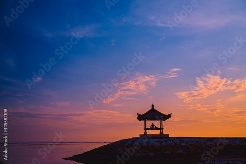 Silhouette of a man sitting in a gazebo on the beach at dawn on Sanur beach, Bali, Indonesia. A man admires the dawn.