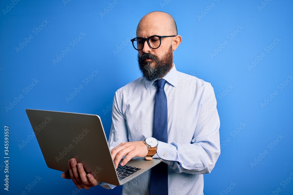 Handsome bald business man with beard working using laptop over blue background with a confident expression on smart face thinking serious