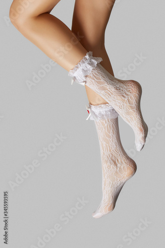 Cropped subject shot of female legs in white lace socks with wide ruffles and silk bows with fabric application on the rims. The photo is made on the gray background. 