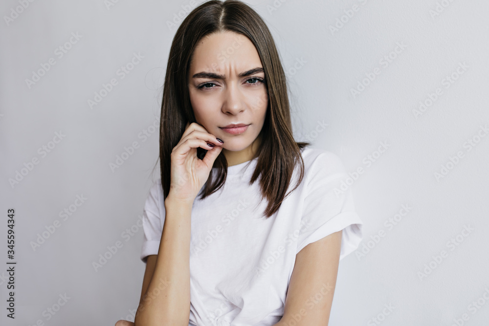 Unhappy charming woman in t-shirt posing in studio. Unpleased dark-haired european girl standing in front of white wall. Unhappy charming woman in t-shirt posing in studio. Unpleased dark-haired european girl standing in front of white wall.